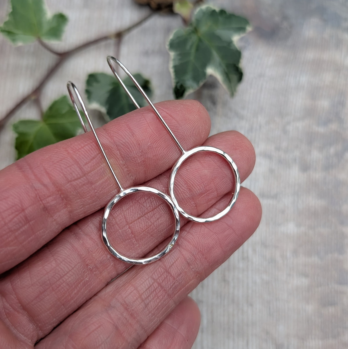 Close up of handmade silver hammered circle long earrings held against a hand, showing the shiny hammered texture, open circle design and long silver ear wires, with ivy leaves in the background.