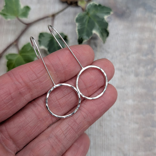 Close up of handmade silver hammered circle long earrings held against a hand, showing the shiny hammered texture, open circle design and long silver ear wires, with ivy leaves in the background.