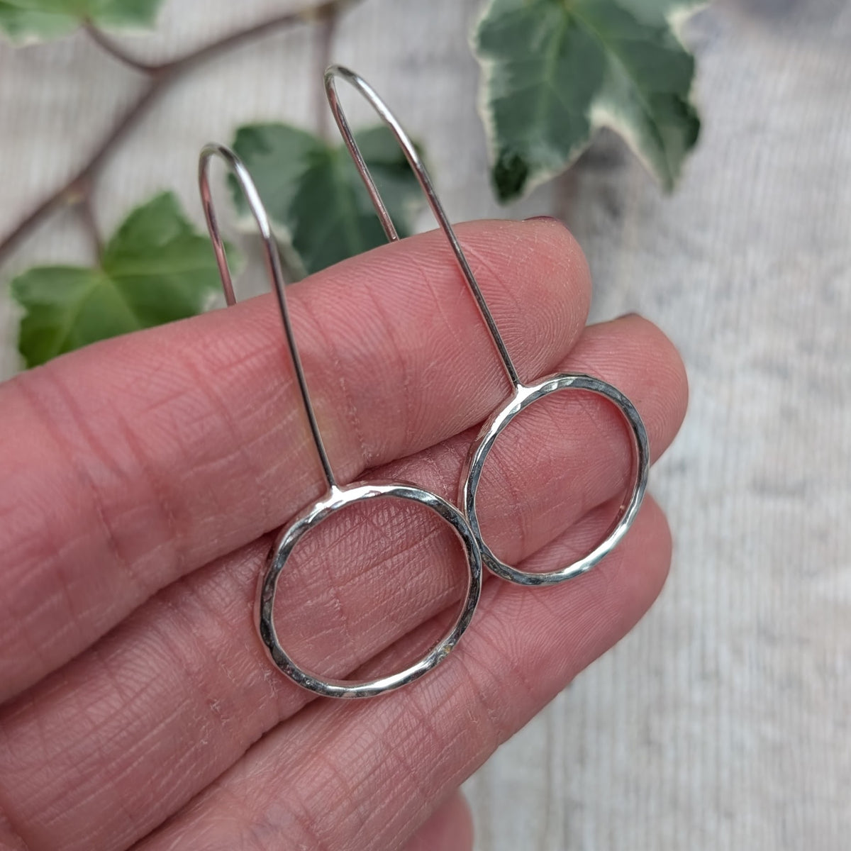 Close up of silver hammered circle long earrings handmade from recycled sterling silver, showing the textured open circles and long silver ear wires resting on a hand with green ivy in the background.