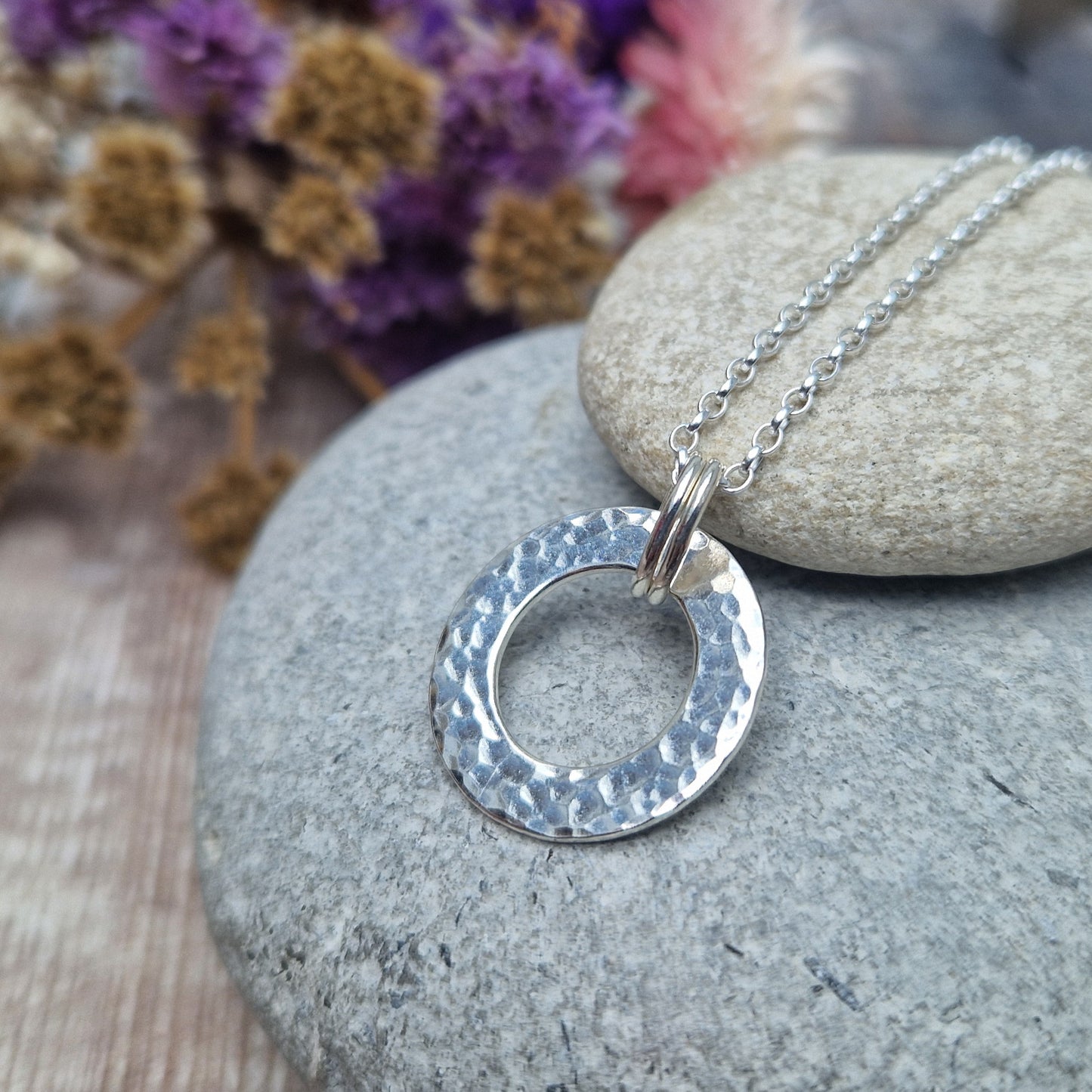 Close-up angled view of a handmade silver hammered circle necklace displayed on smooth stones, showing the texture and shine of the silver.