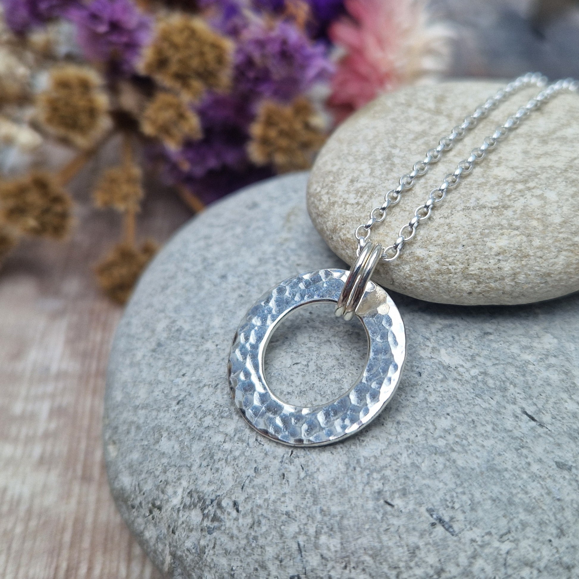 Close-up angled view of a handmade silver hammered circle necklace displayed on smooth stones, showing the texture and shine of the silver.