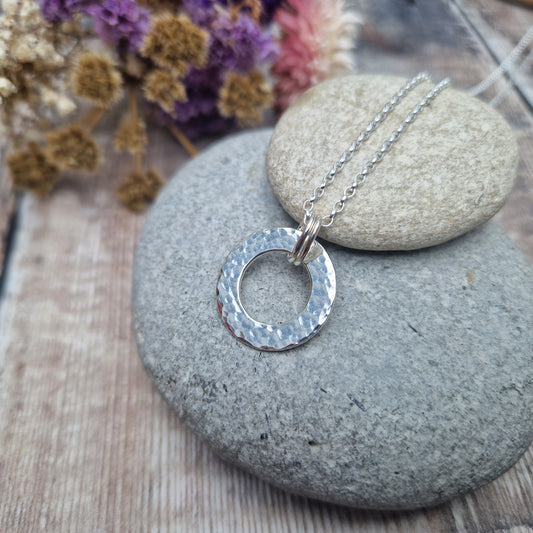 Close-up of a handmade silver hammered circle necklace displayed on two stacked smooth stones, with a blurred background of dried flowers.