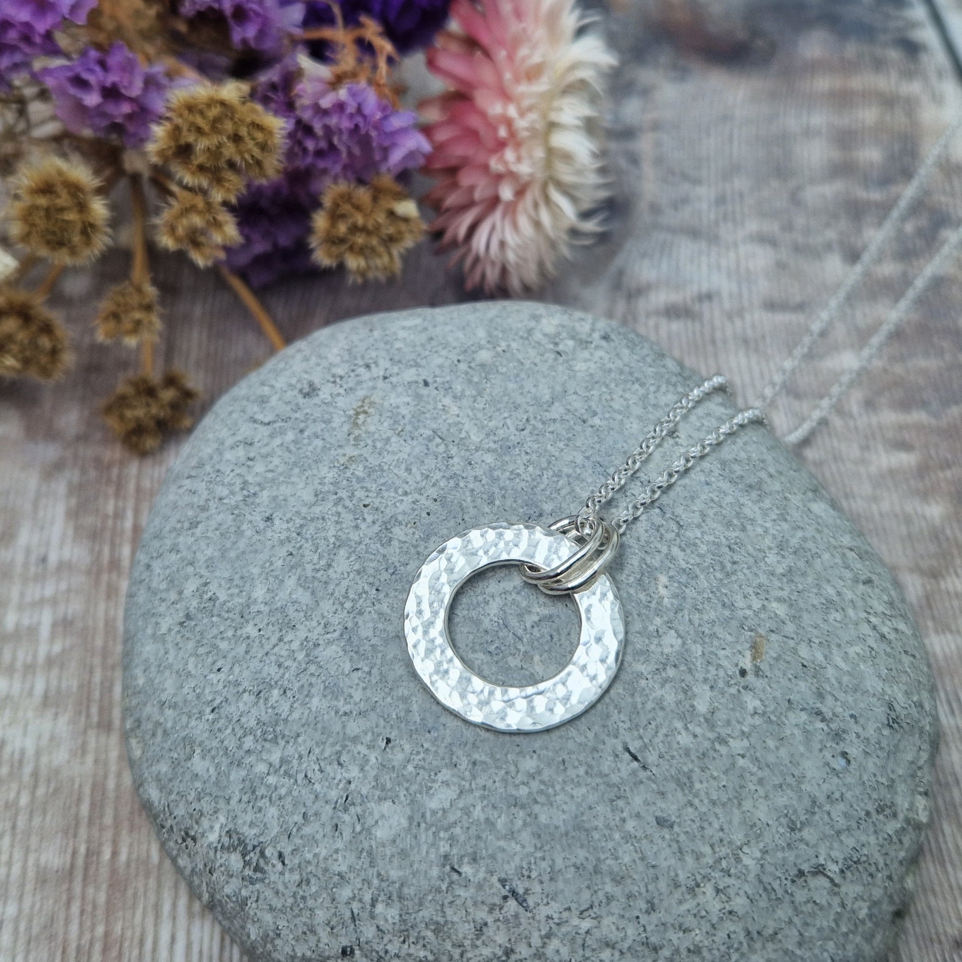 Handmade silver hammered circle necklace with three small rings on the pendant, resting on a grey stone, with dried flowers in the background.