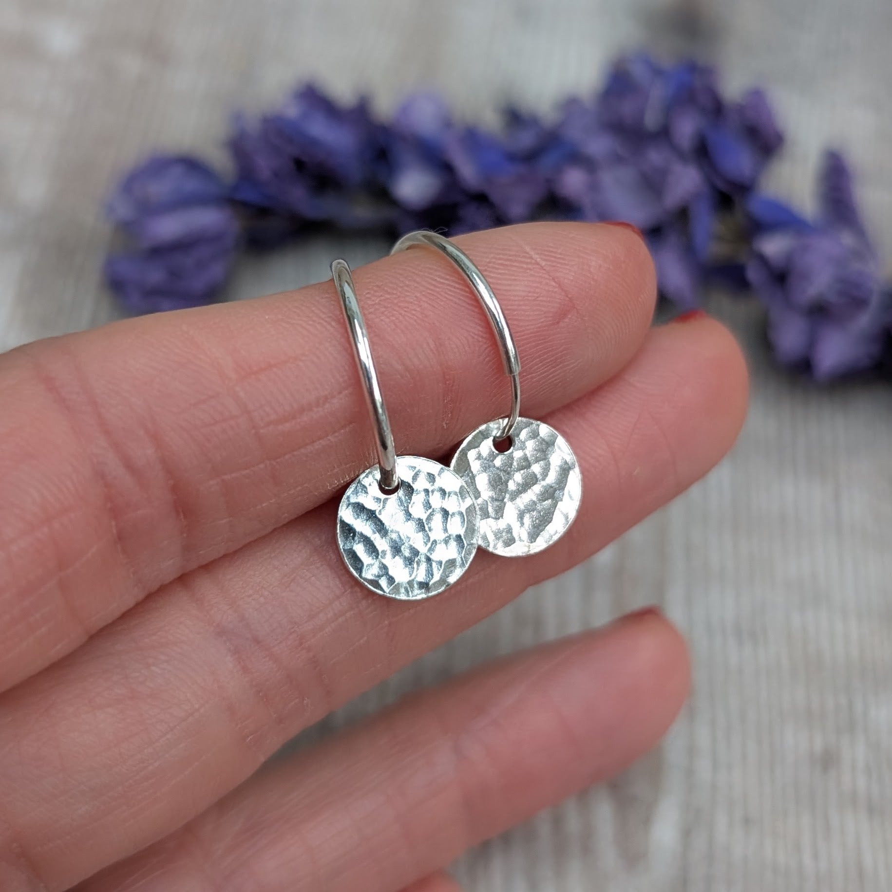 Hand holding a pair of Silver Hammered Disc Hoop Earrings, showing the small circular hammered discs hanging from simple silver hoops with a blurred lavender background.