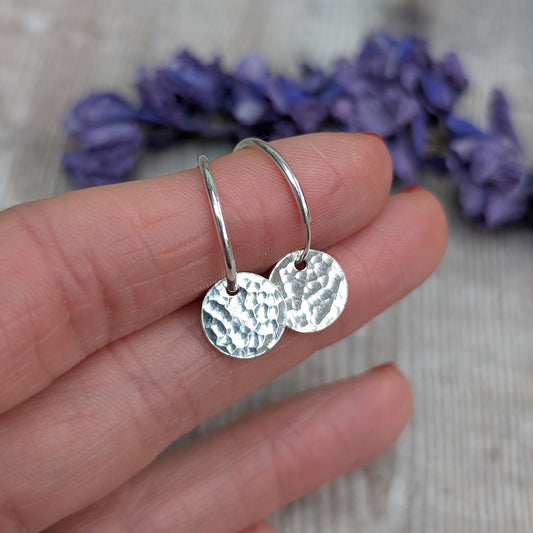 Hand holding a pair of Silver Hammered Disc Hoop Earrings, showing the small circular hammered discs hanging from simple silver hoops with a blurred lavender background.