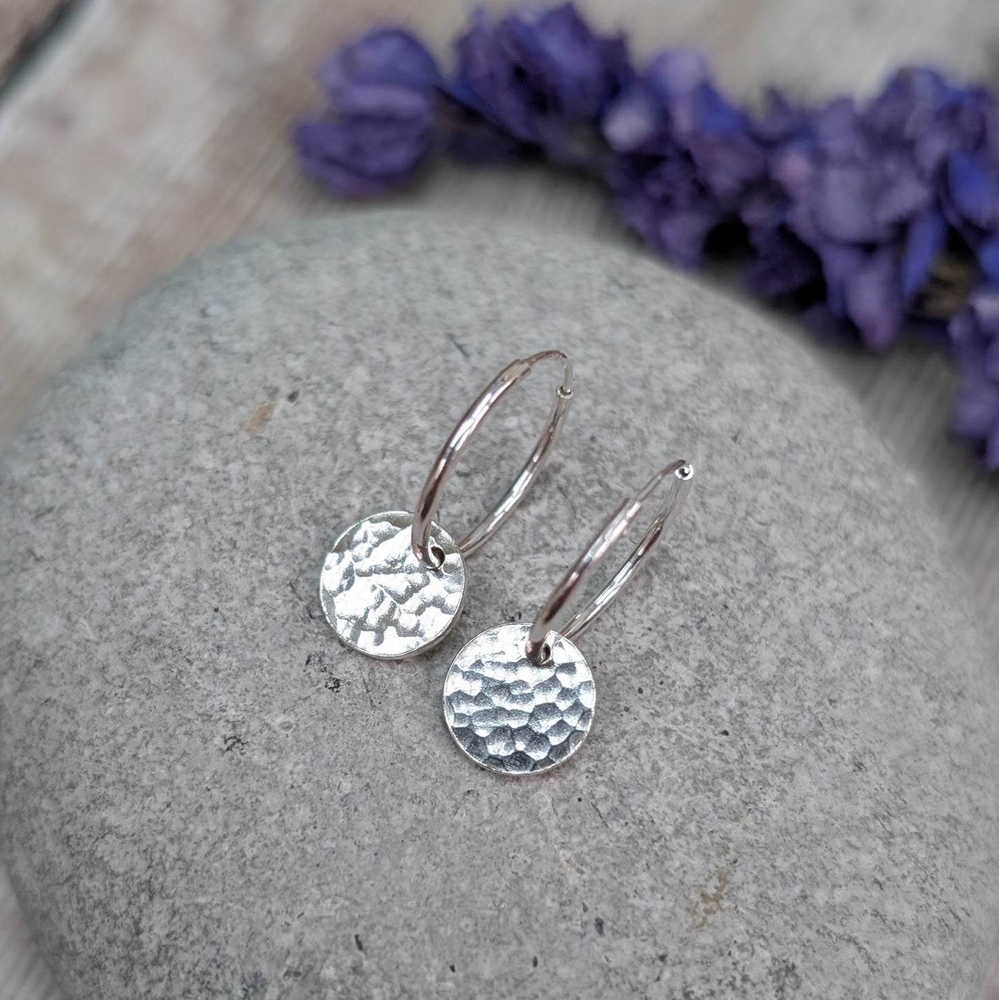 Silver Hammered Disc Hoop Earrings resting on a smooth grey stone, with dried purple flowers softly blurred in the background.