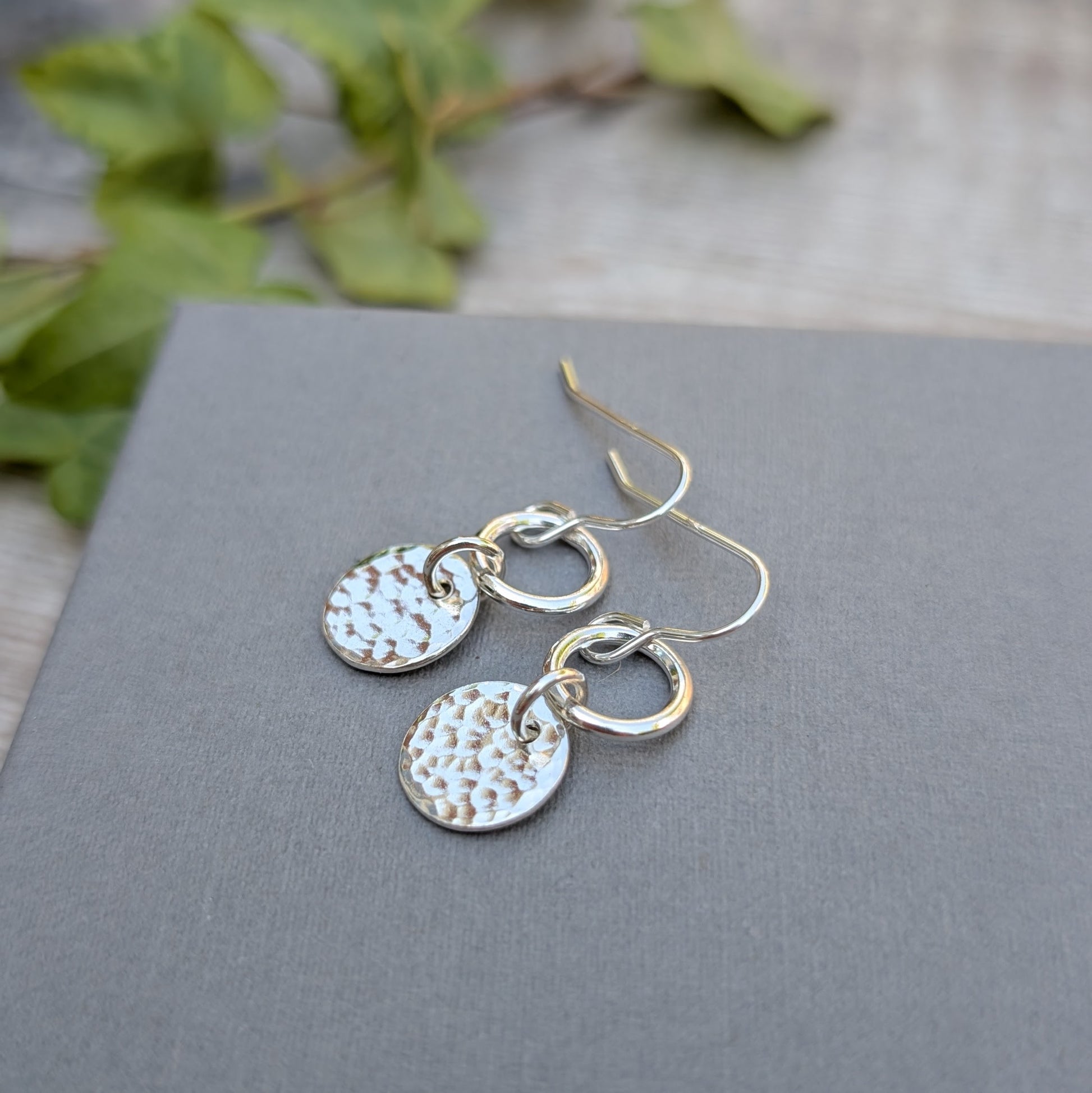 Silver hammered disc and circle earrings handmade from recycled Sterling Silver displayed on a grey card with a rustic wooden background and ivy detail.