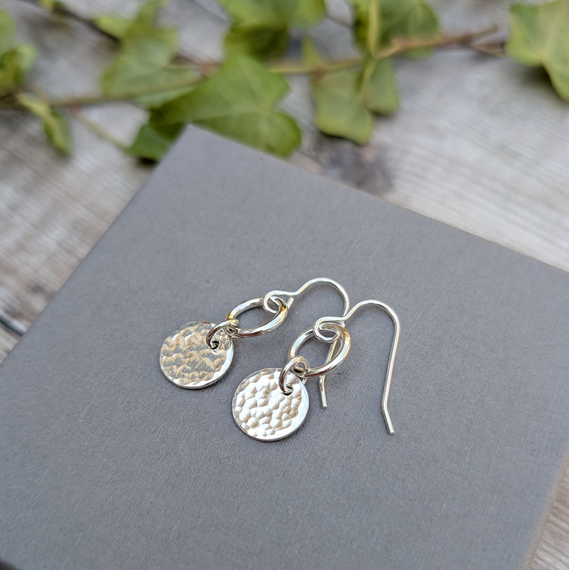 Silver hammered disc and circle earrings handmade from recycled Sterling Silver, shown on a grey display card with a soft ivy background.