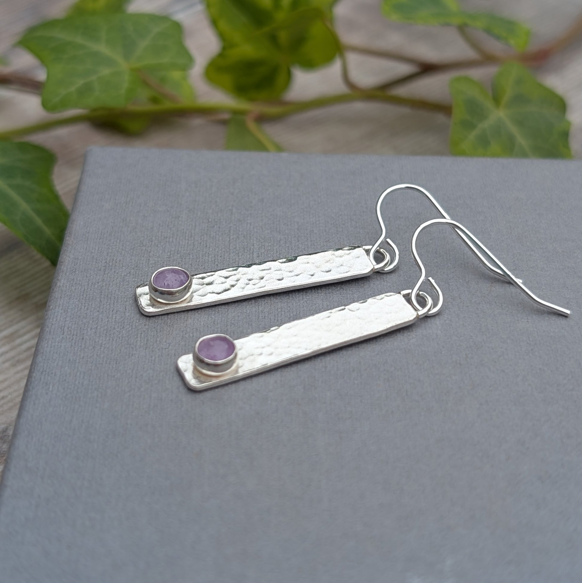 Silver Hammered Rectangle Earrings with Lavender Amethyst laid on a grey surface with greenery in the background