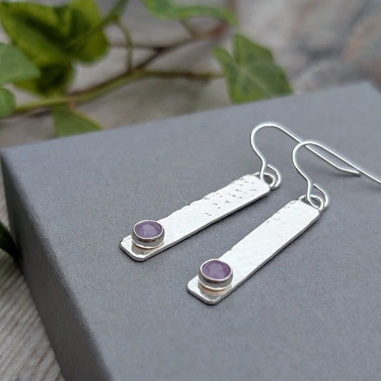 Silver Hammered Rectangle Earrings with Lavender Amethyst resting on a grey box with green leaves in the background