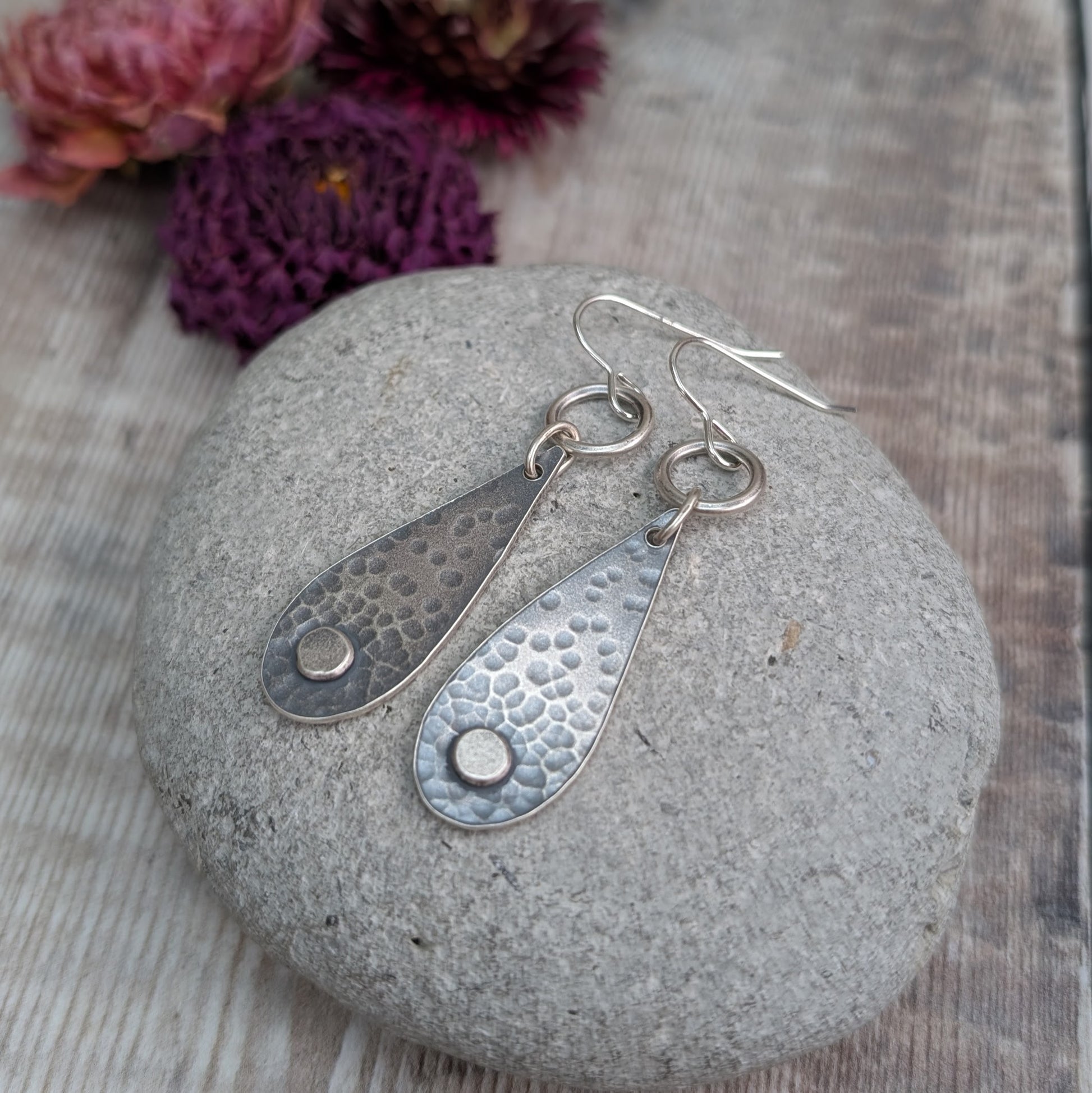 Each earring  teardrop in shape with oxidised hammered texture, accentuated around a small, raised pebble at the wider part of the teardrop. Suspended from earring wire via an open circle link so that the earring displays front facing when worn.