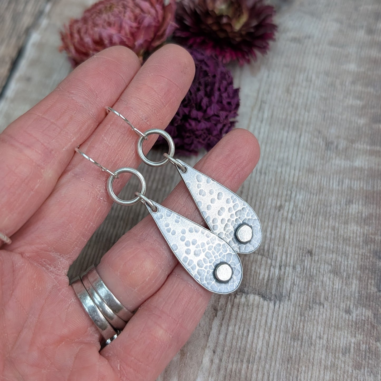 Each earring  teardrop in shape with oxidised hammered texture, accentuated around a small, raised pebble at the wider part of the teardrop. Suspended from earring wire via an open circle link so that the earring displays front facing when worn.