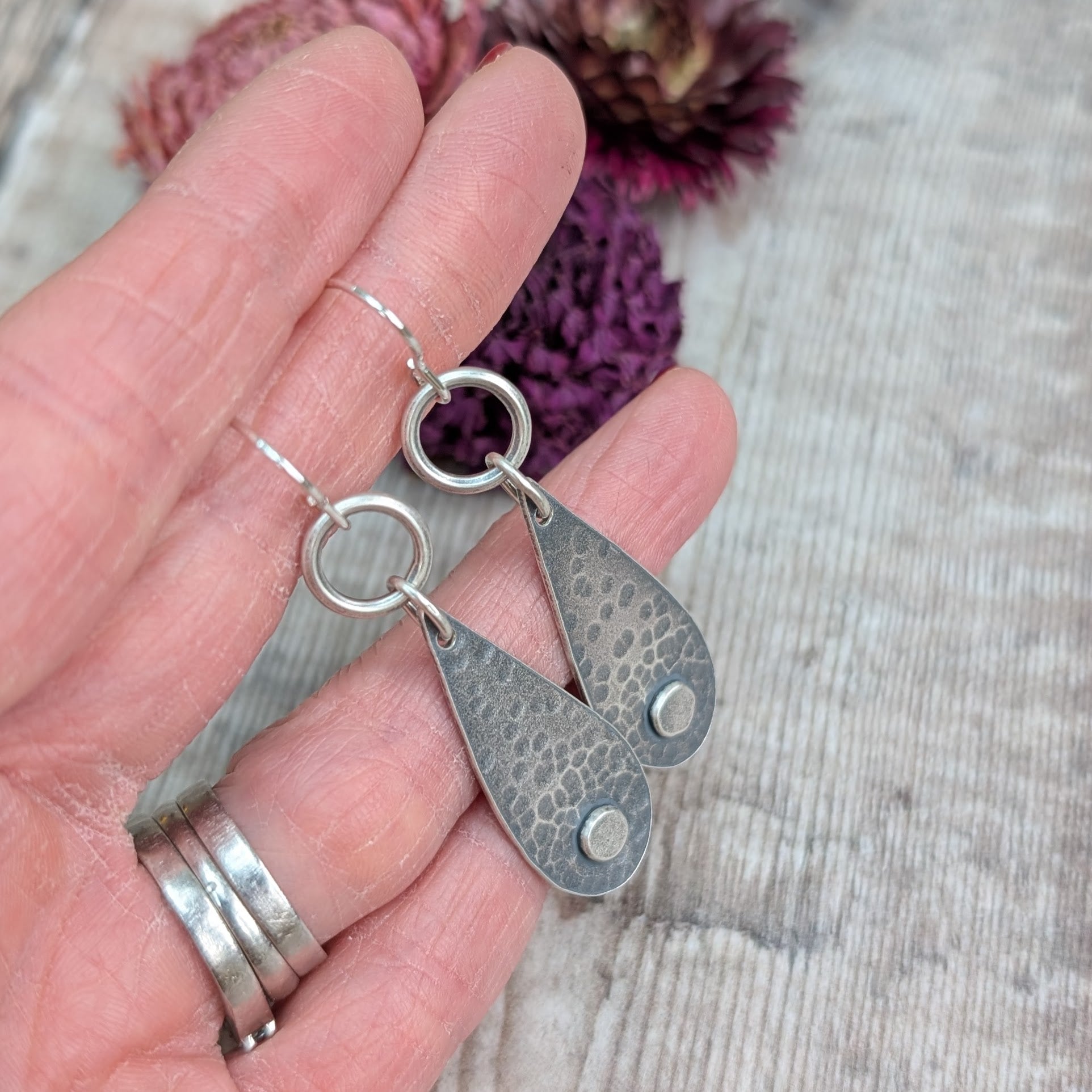 Each earring  teardrop in shape with oxidised hammered texture, accentuated around a small, raised pebble at the wider part of the teardrop. Suspended from earring wire via an open circle link so that the earring displays front facing when worn.