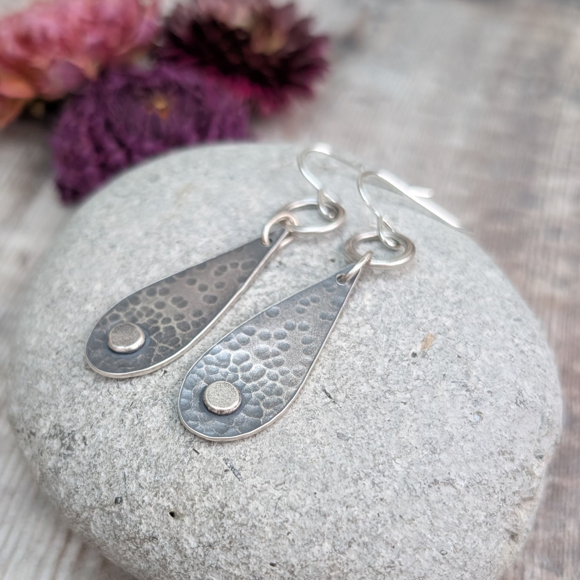 Each earring  teardrop in shape with oxidised hammered texture, accentuated around a small, raised pebble at the wider part of the teardrop. Suspended from earring wire via an open circle link so that the earring displays front facing when worn.