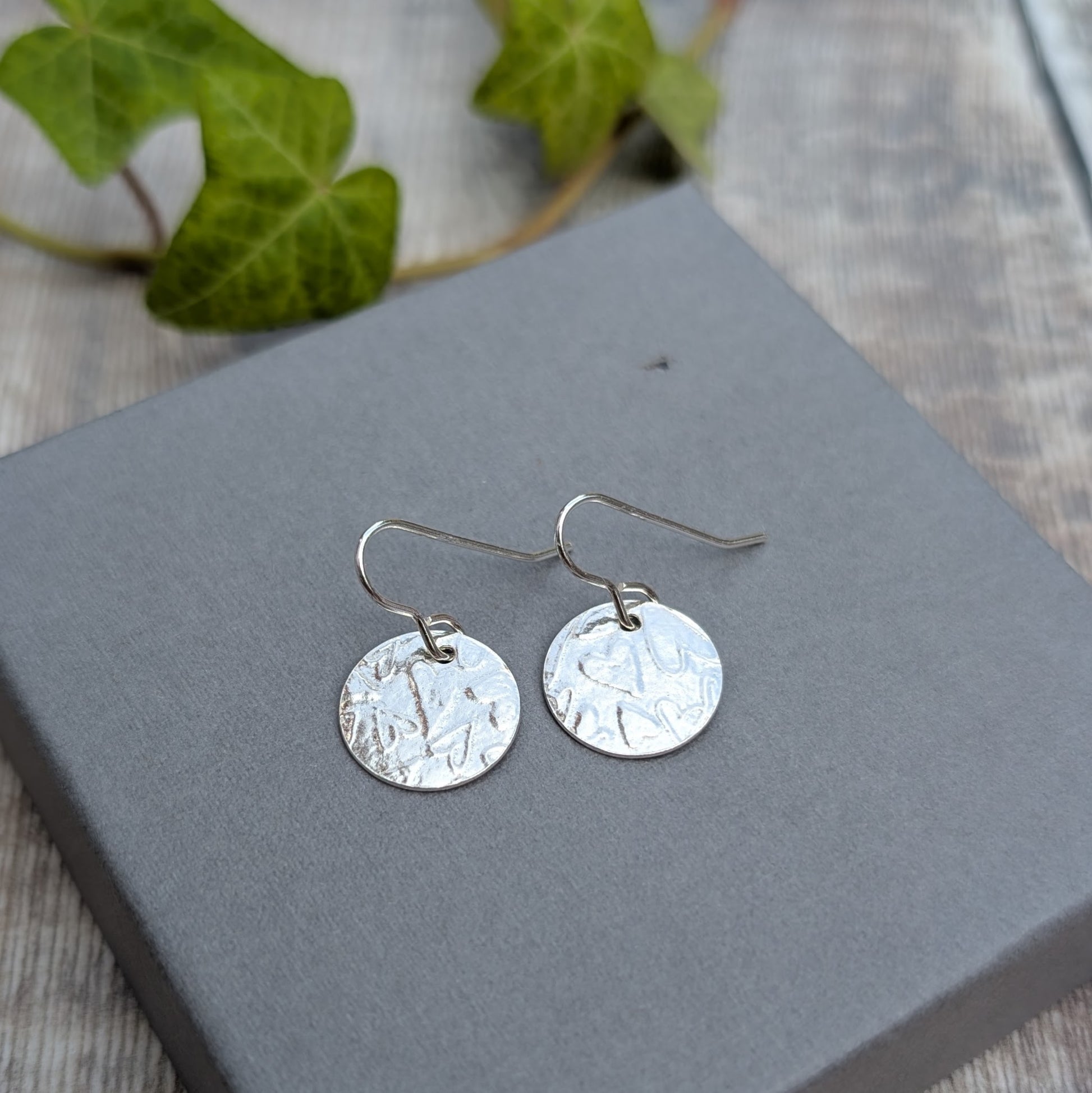 Silver heart pattern disc earrings handmade from recycled Sterling Silver, with textured round discs and silver ear wires, displayed on a grey background.