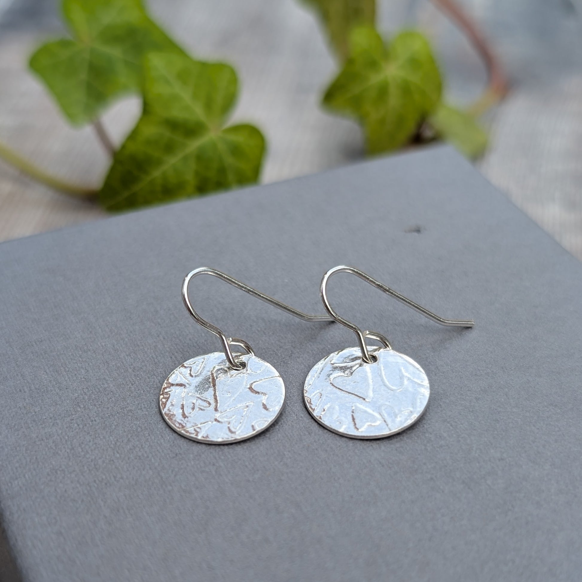 Silver heart pattern disc earrings handmade from recycled Sterling Silver, shown on a grey background with green ivy.