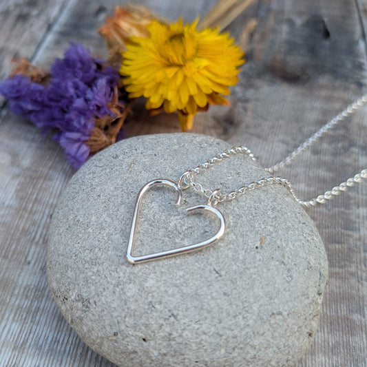 Silver heart ring keeper necklace displayed on a smooth grey stone, with yellow and purple flowers in the background.