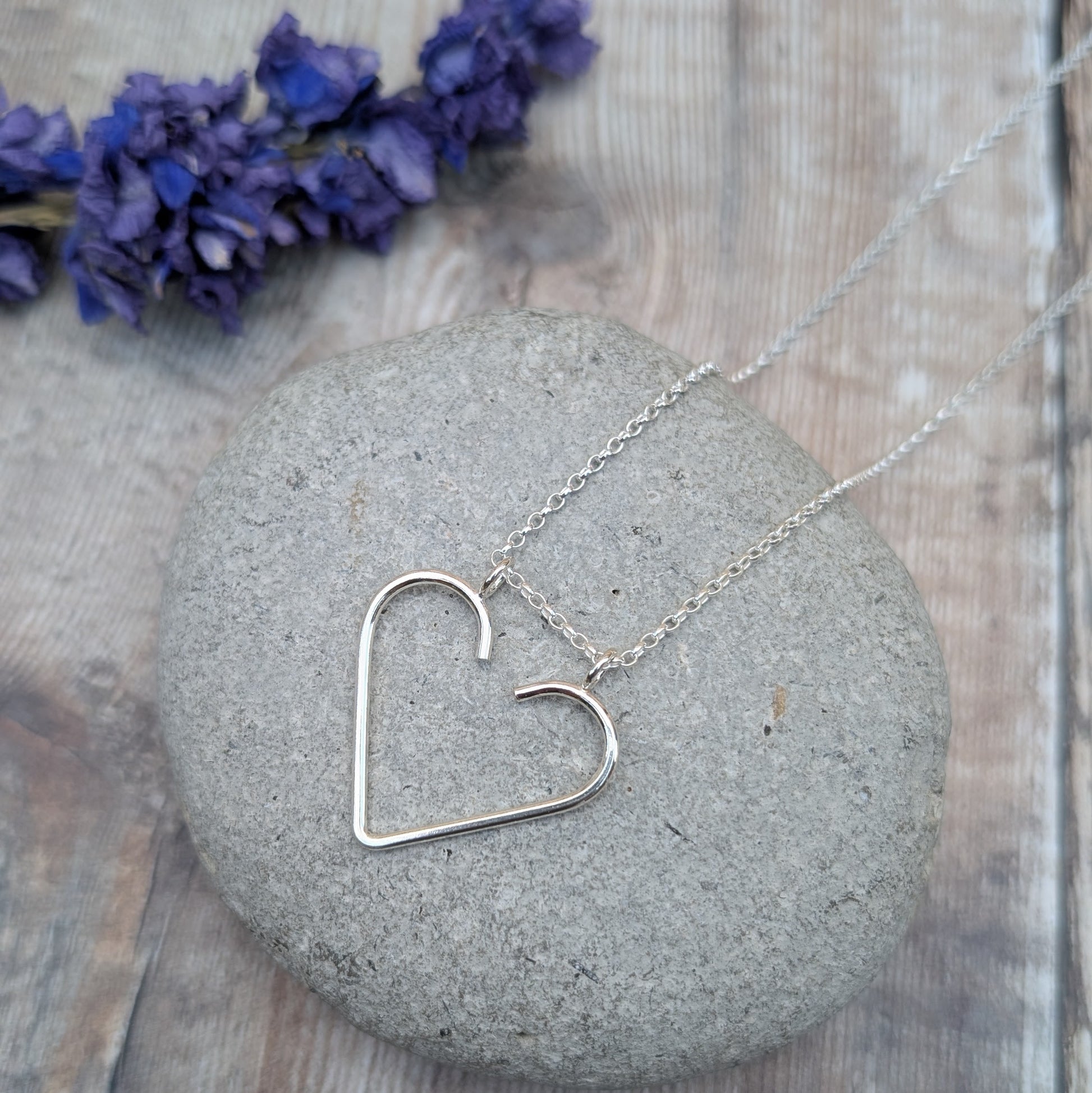 Silver heart ring keeper necklace placed on a grey stone with purple flowers in the background.