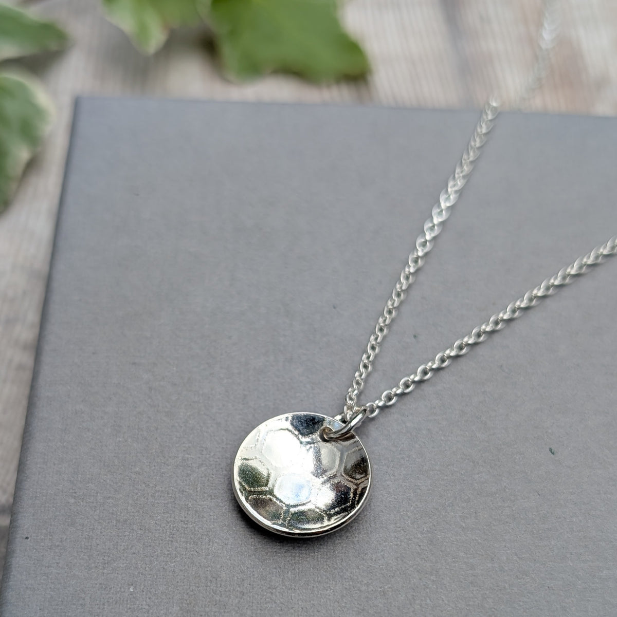 Silver honeycomb disc necklace displayed on a dark grey background, showing the round curved pendant and honeycomb texture.