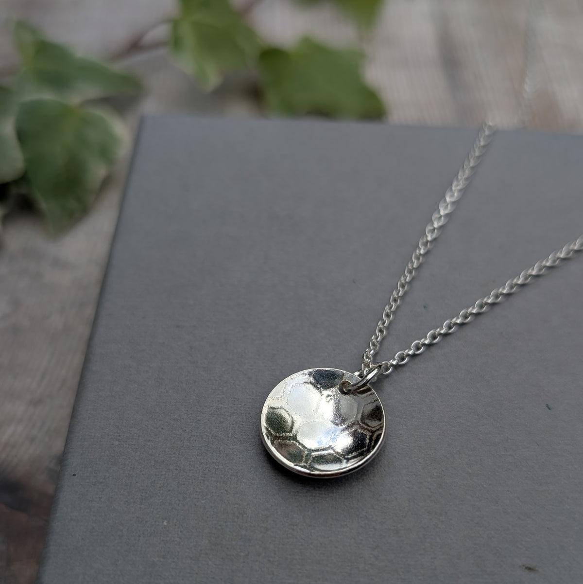 Silver honeycomb disc necklace handmade from recycled Sterling Silver, displayed on a dark grey background with a leafy backdrop.