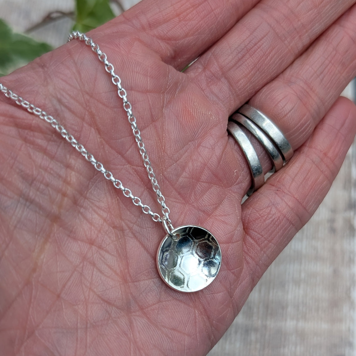 Silver honeycomb disc necklace held in a hand, showing the size and handmade honeycomb patterned pendant.