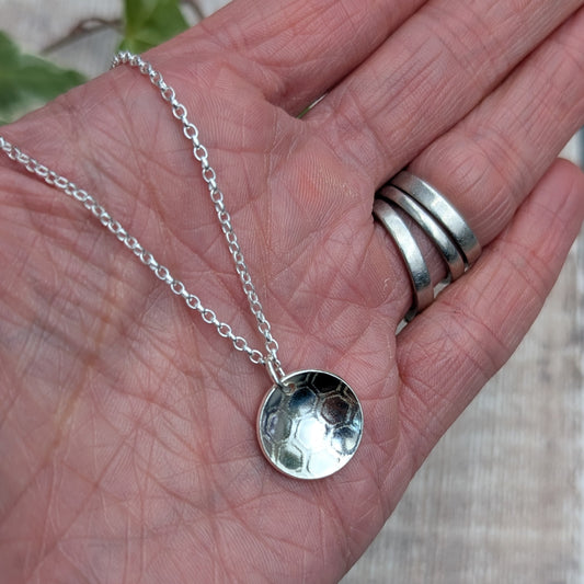 Silver honeycomb disc necklace held in a hand, showing the size and handmade honeycomb patterned pendant.