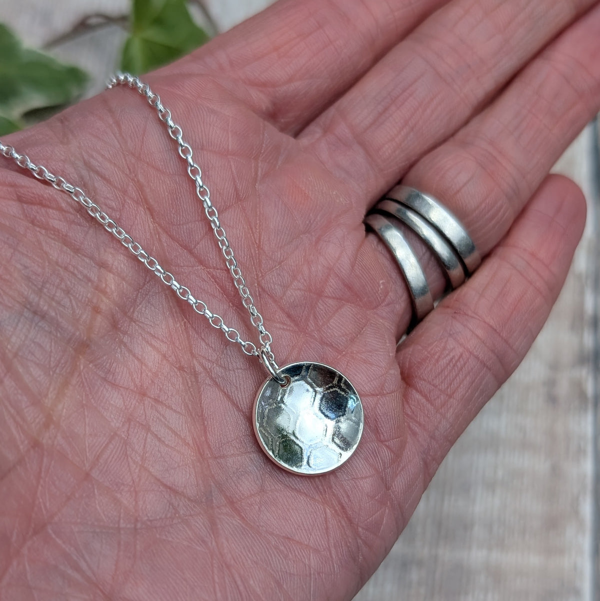 Close up of a silver honeycomb disc necklace held in a hand, showing the detailed pattern pressed into the recycled Sterling Silver.