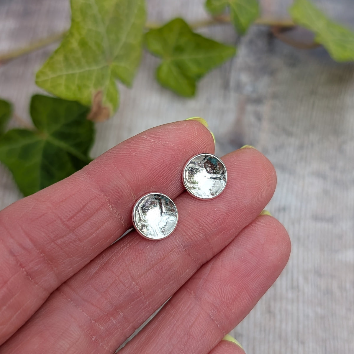 Silver honeycomb disc stud earrings handmade from recycled Sterling Silver, shown resting on a hand with ivy in the background.