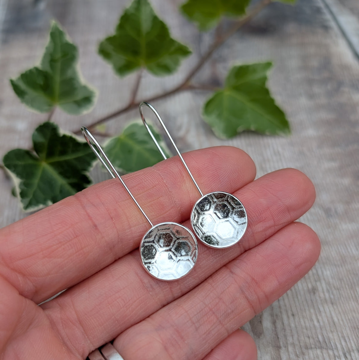 Silver honeycomb long disc earrings handmade from recycled Sterling Silver, shown resting on a hand with green ivy in the background.