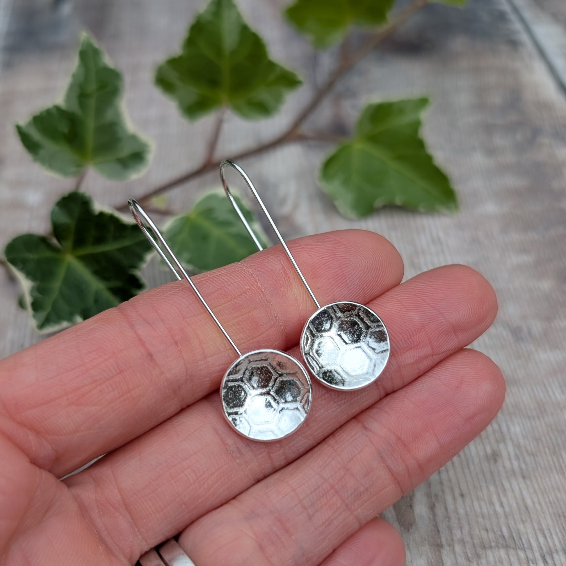Silver honeycomb long disc earrings handmade from recycled Sterling Silver, shown resting on a hand with green ivy in the background.