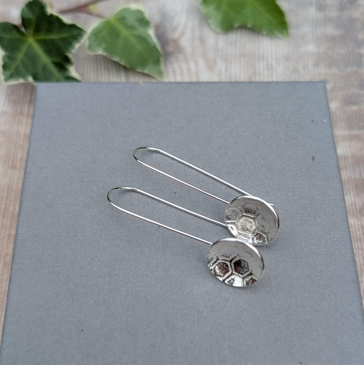 Handmade silver honeycomb long disc earrings lying on a grey display surface with green ivy in the background.