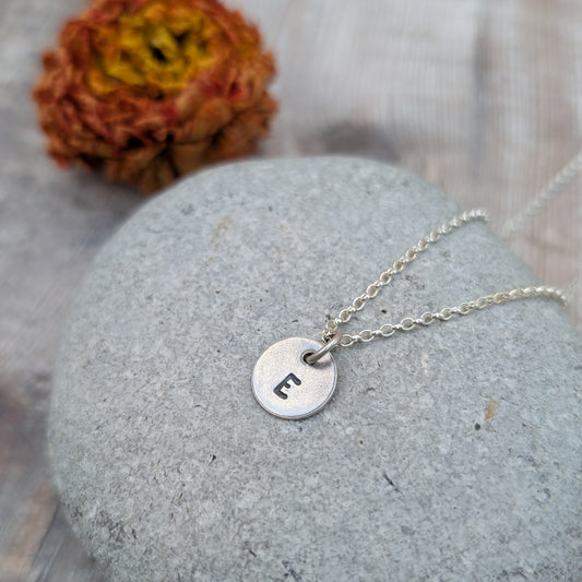 A silver initial disc necklace with the letter "E" stamped on the pendant, displayed on a grey stone background.