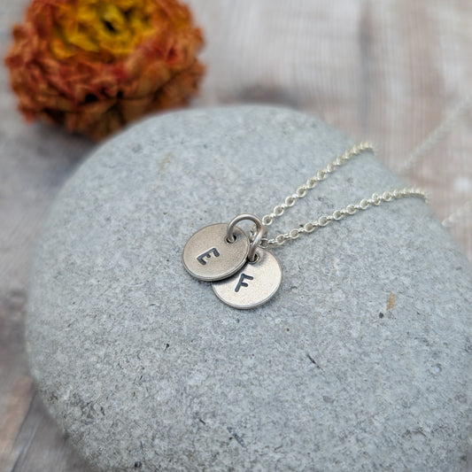 A silver necklace with two stamped initial discs, letters "E" and "F," resting on a grey stone.