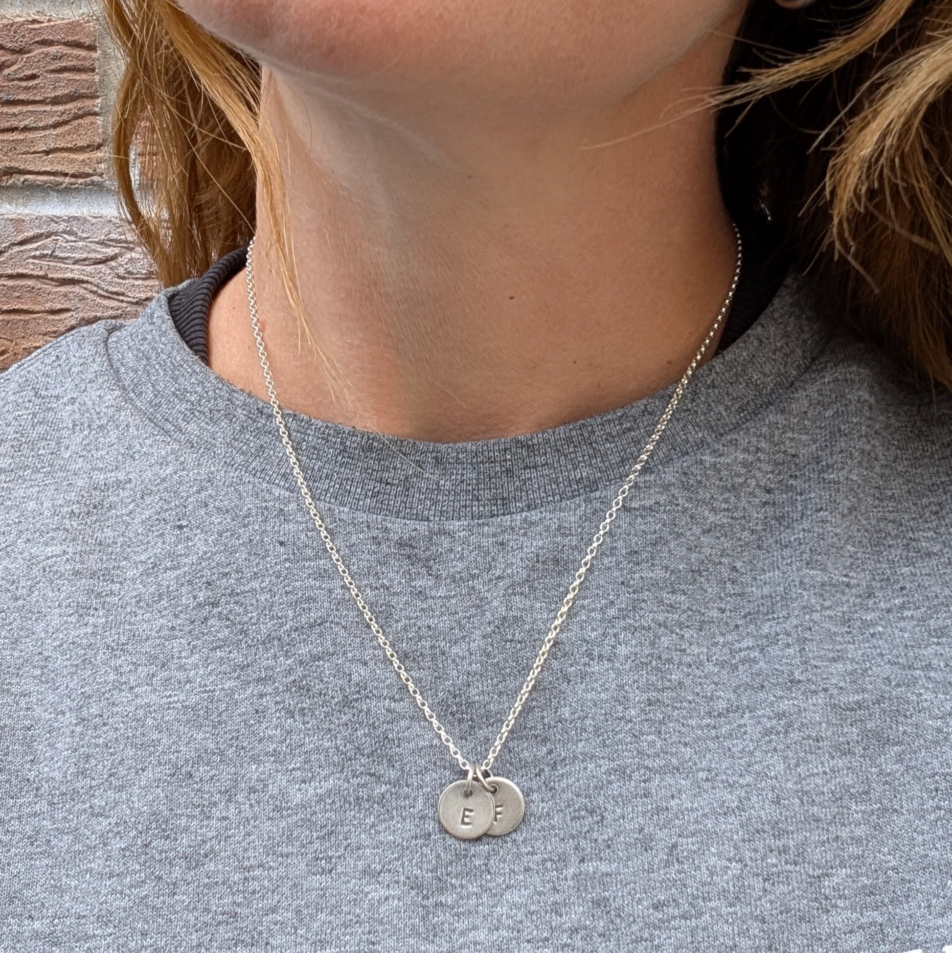 A woman wearing a silver initial disc necklace with two stamped pendants, "E" and "F," shown against a grey top.