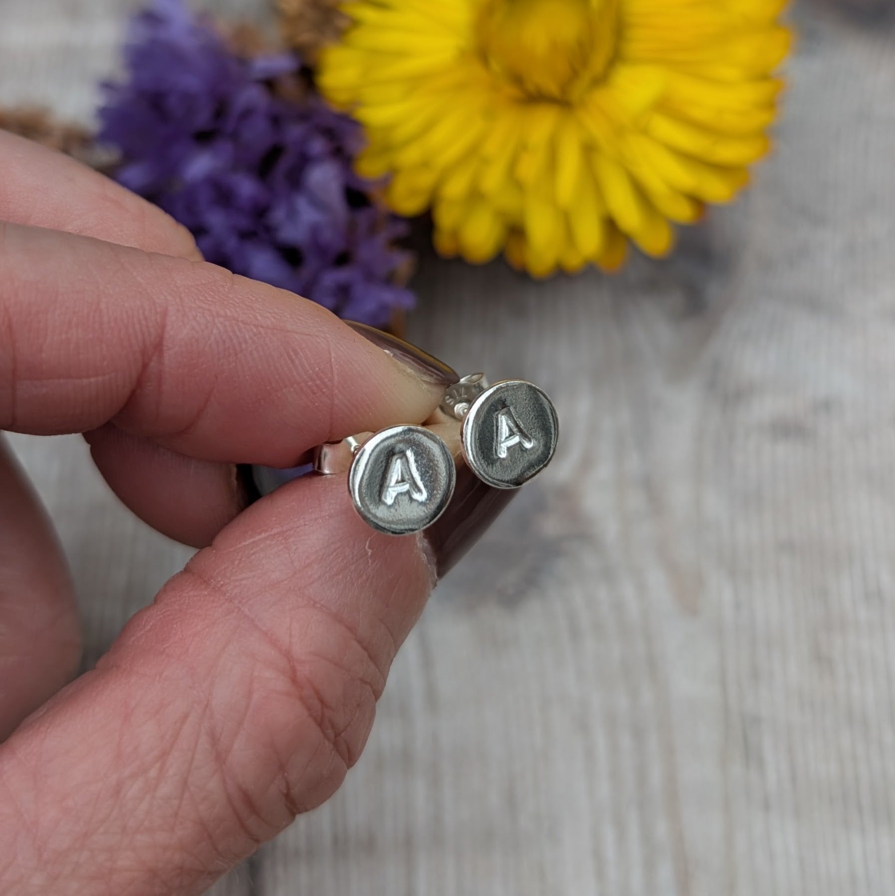 Pair of silver initial stud earrings with the letter “A,” held between fingers.