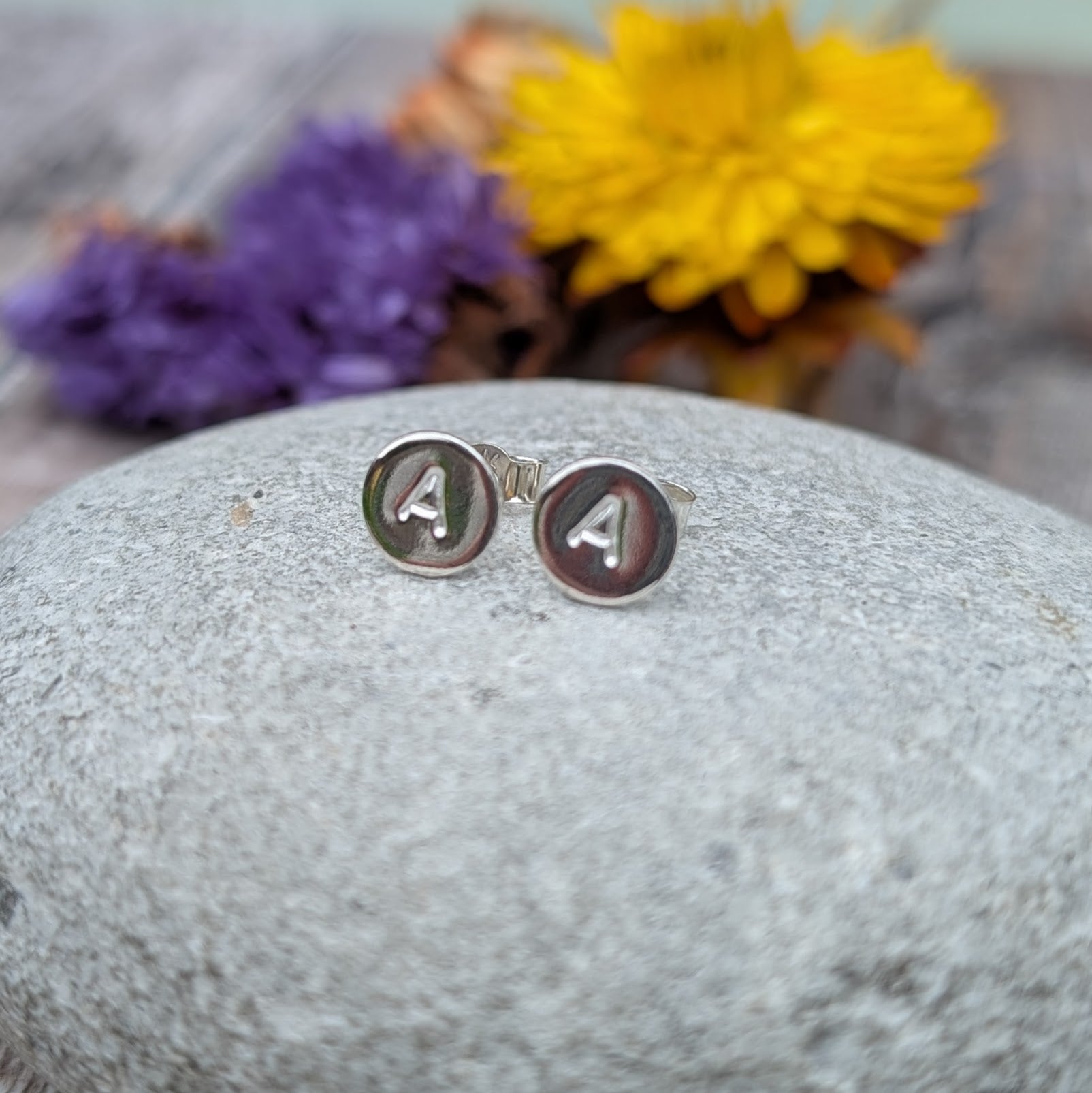 Silver initial stud earrings displayed on a smooth grey stone with blurred yellow and purple flowers.