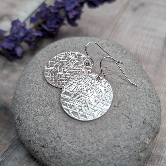A pair of silver large textured hammered disc earrings resting on a smooth grey stone with purple flowers in the background.