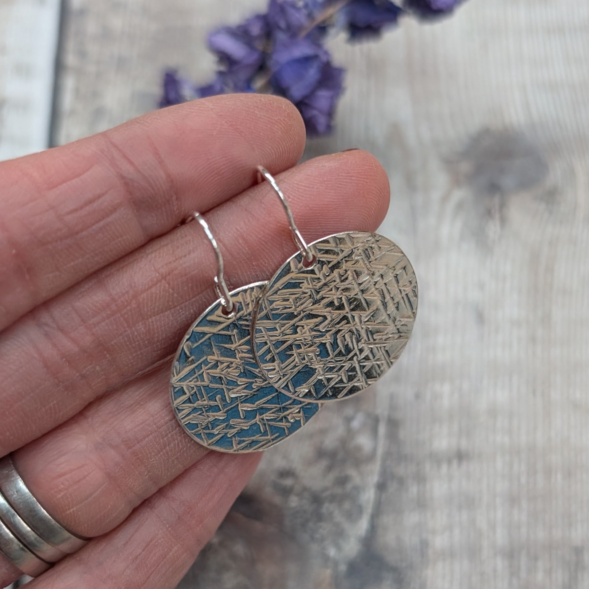 Close-up of a hand holding silver large textured hammered disc earrings showing the detailed cross-hatched surface.