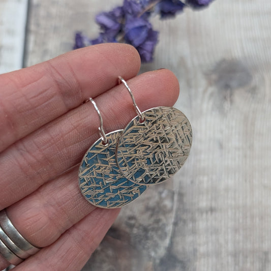 Close-up of a hand holding silver large textured hammered disc earrings showing the detailed cross-hatched surface.