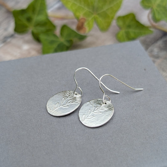 Silver leafy vine oval disc earrings handmade from recycled Sterling Silver, displayed on a grey background with green ivy behind.