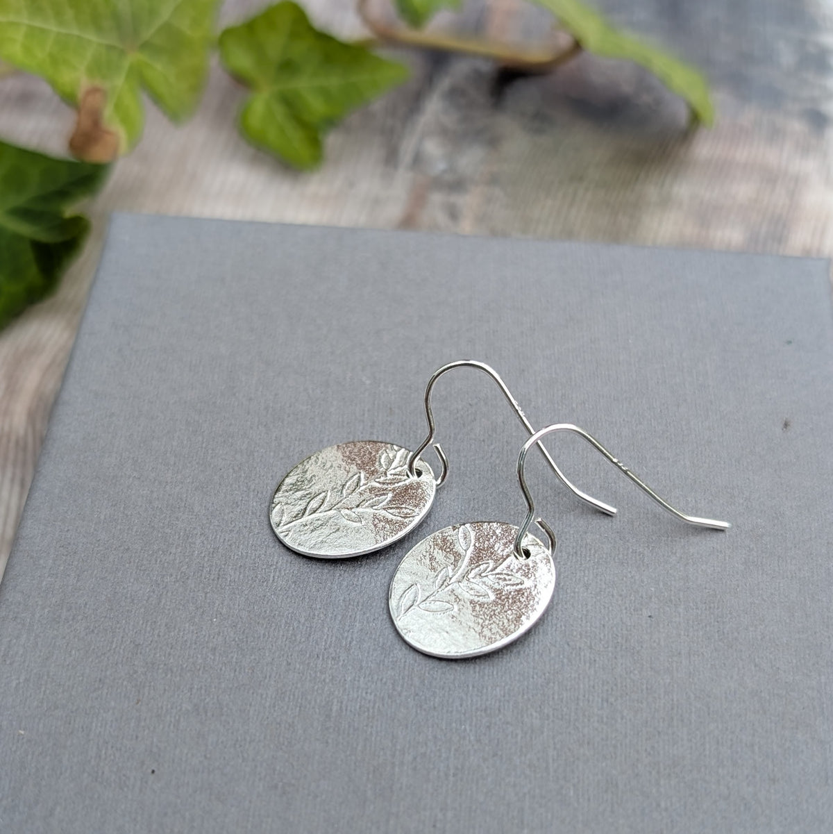 Silver leafy vine oval disc earrings with a pressed leaf and branch pattern, shown resting on a grey background.