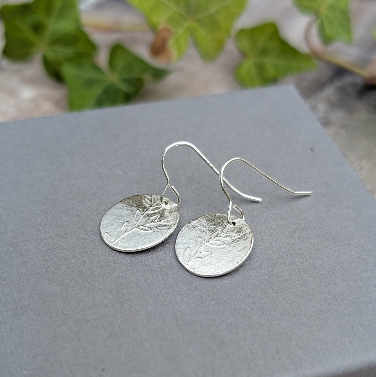 Close up of silver leafy vine oval disc earrings showing the textured silver surface and leafy pattern pressed into the oval discs.