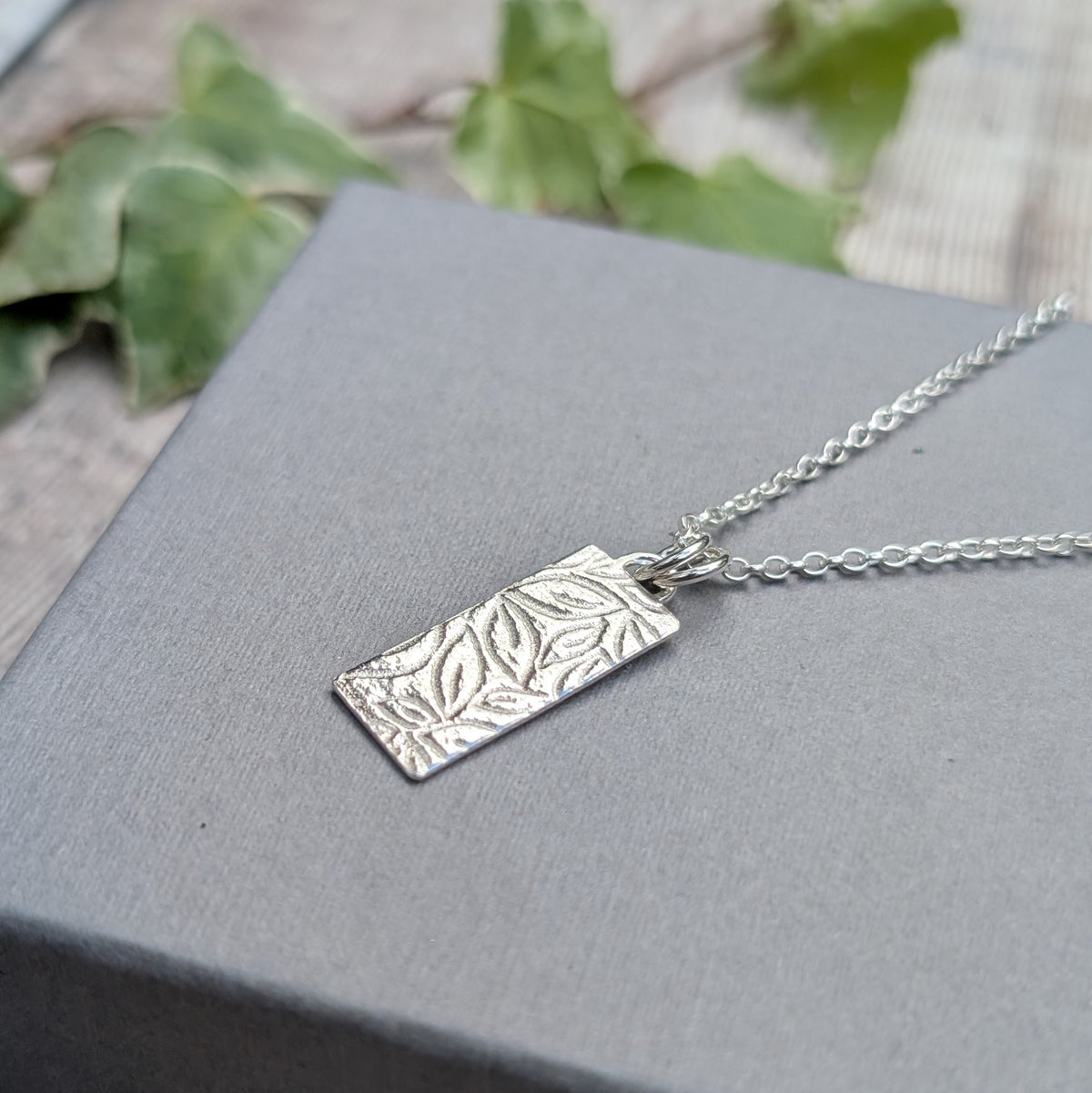 Silver leaves rectangle necklace handmade from recycled Sterling Silver with a textured leaf pattern, displayed on a grey jewellery box with green leaves in the background.