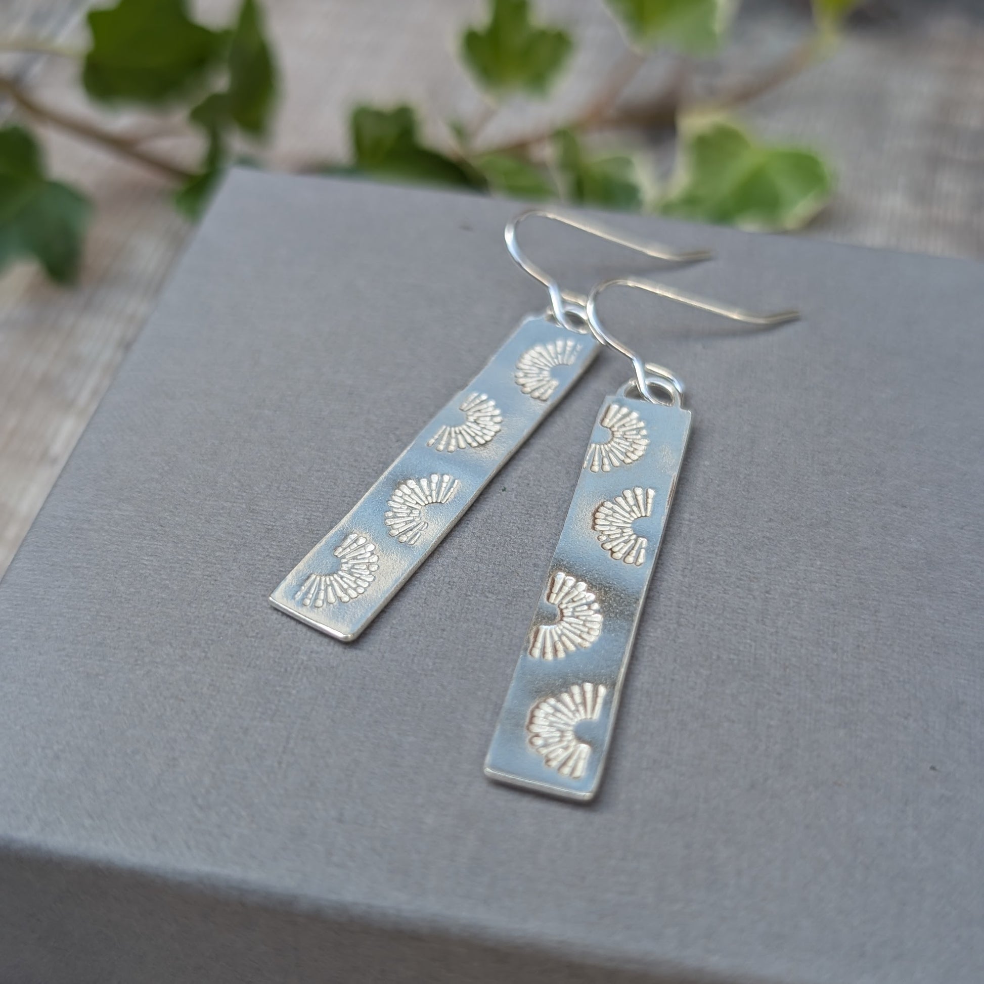 Close up of silver long rectangle sunrise earrings on a grey box, showing the repeating hand stamped sunrise design and silver ear wires.