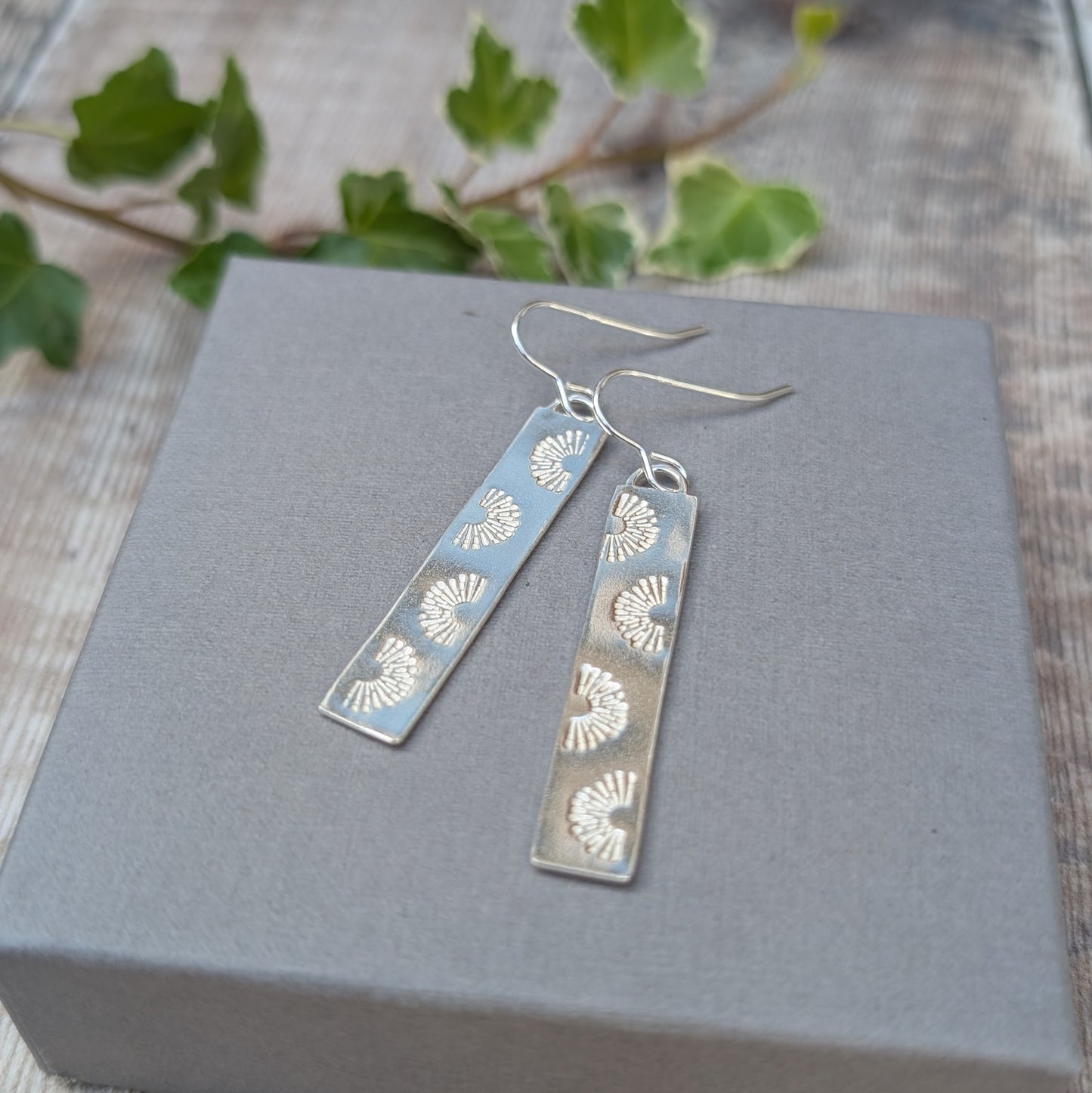 Silver long rectangle sunrise earrings handmade from recycled Sterling Silver, displayed on a grey box with green leaves in the background.