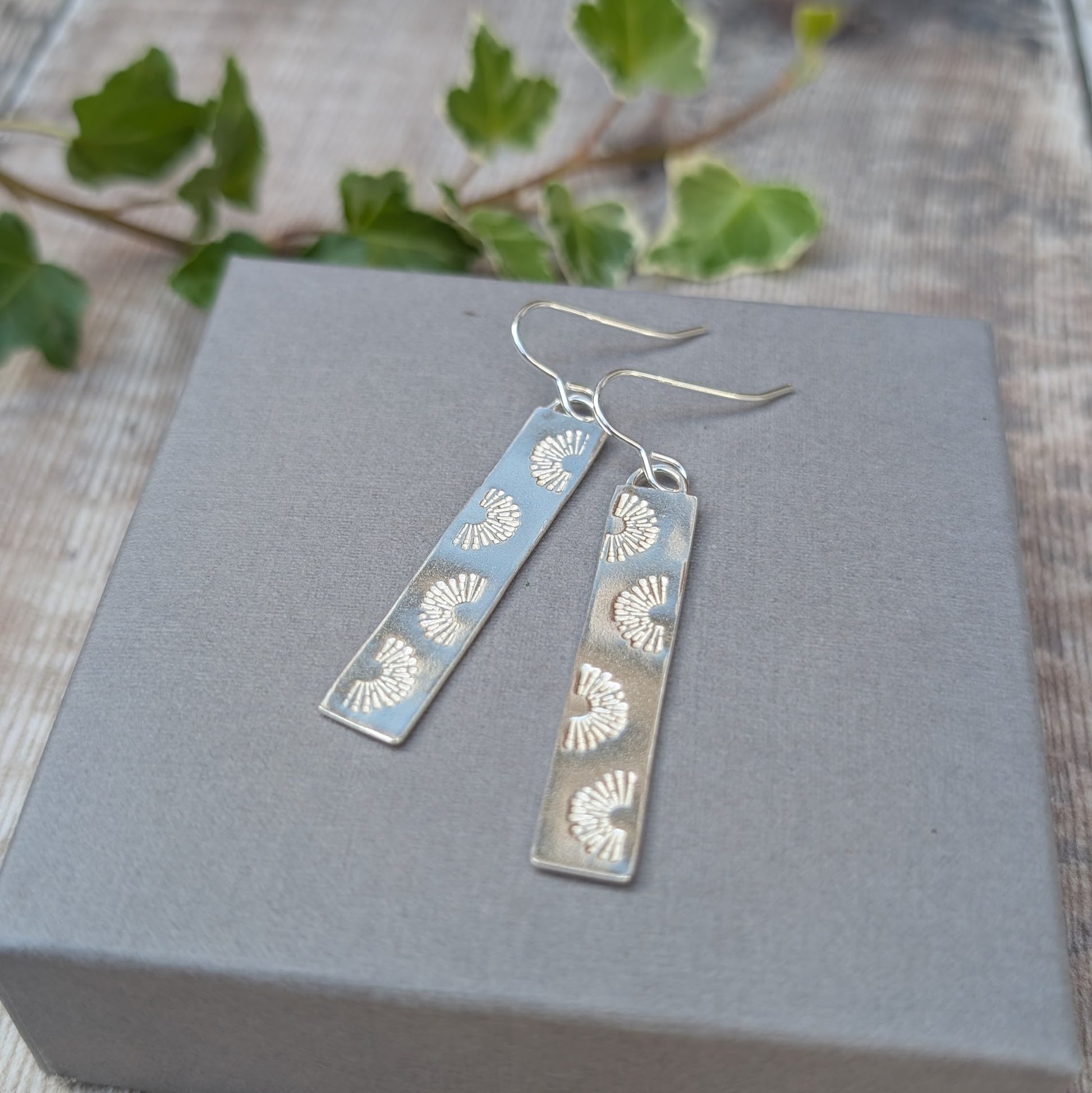 Silver long rectangle sunrise earrings handmade from recycled Sterling Silver, displayed on a grey box with green leaves in the background.