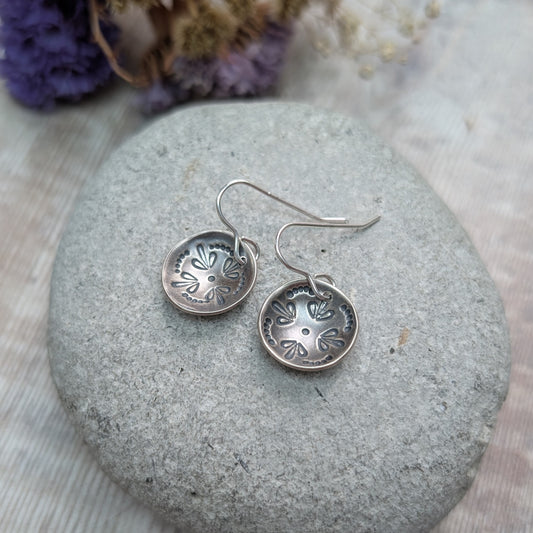 Close-up of a pair of oxidised Silver Mandala Dome Disc Earrings resting on a smooth grey stone, showing the detailed stamped mandala design in the domed surface.