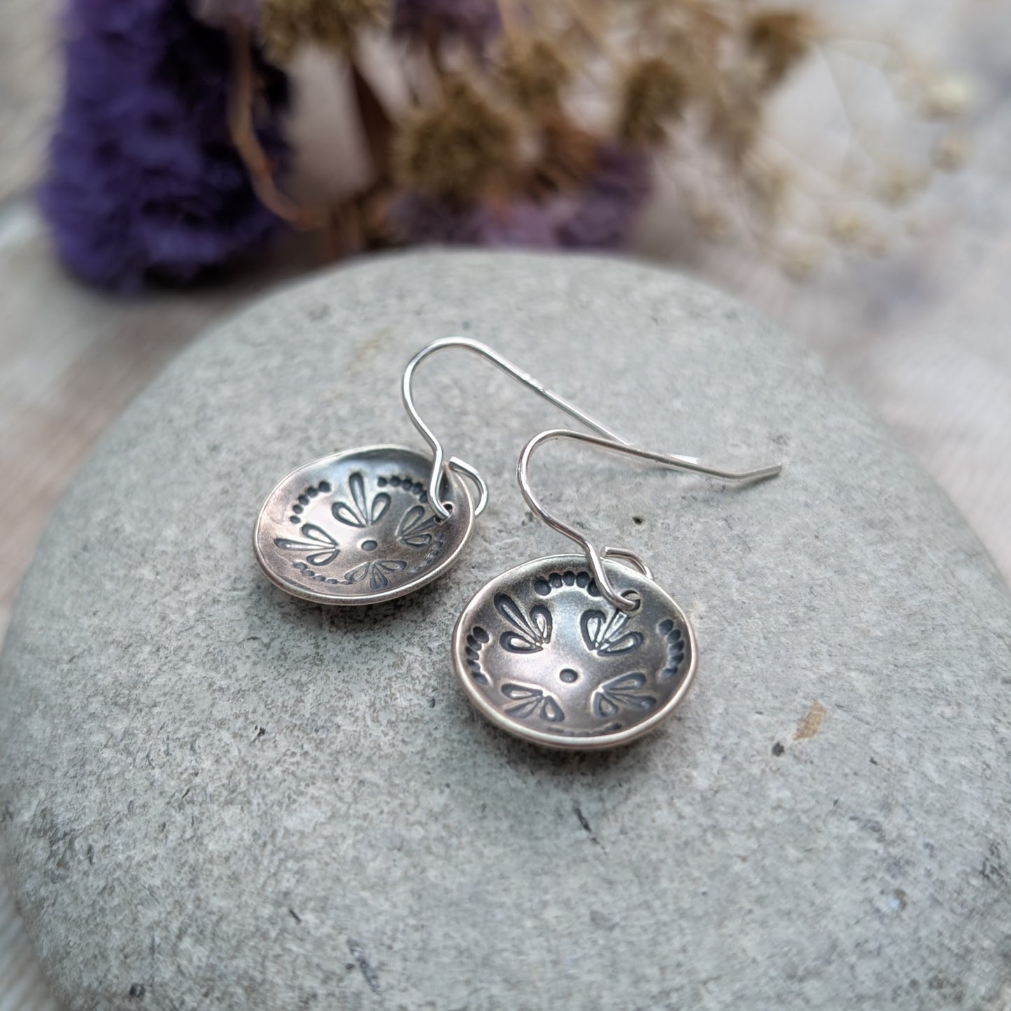 Close-up of a pair of oxidised Silver Mandala Dome Disc Earrings resting on a smooth grey stone, showing the detailed stamped mandala design in the domed surface.