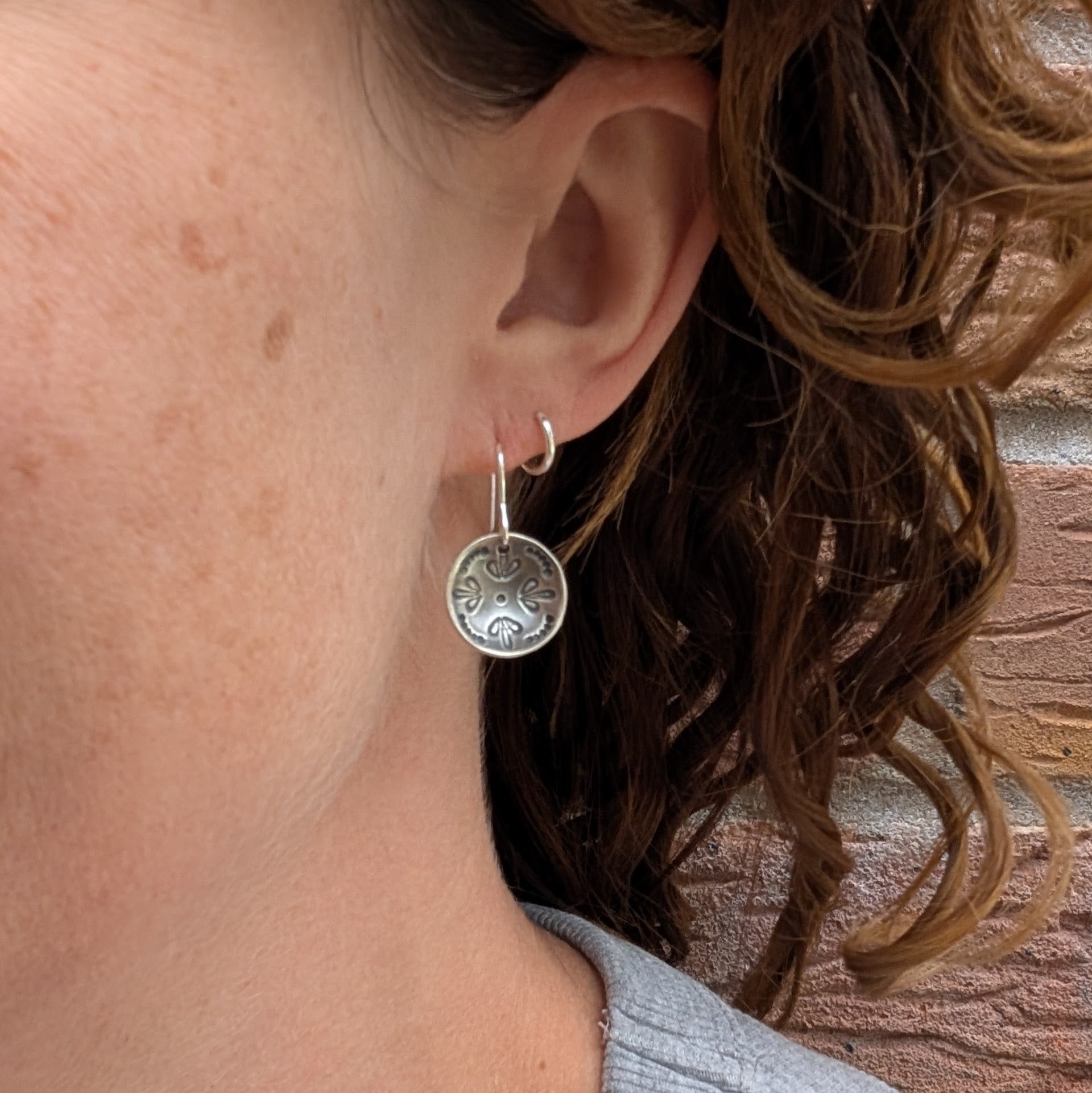 A woman wearing one of the Silver Mandala Dome Disc Earrings, showing how it hangs just below the earlobe with its subtle dome shape and darkened detail.