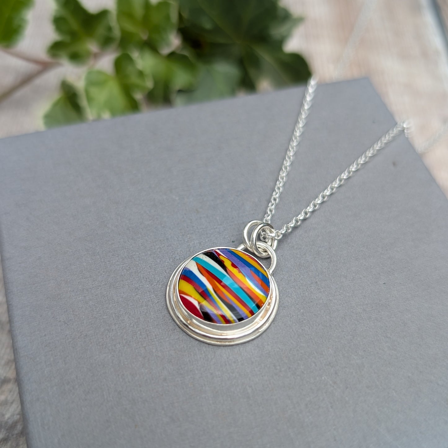 Close up of the Silver Multicoloured Surfite Round Necklace with a bright striped Surfite stone in shades of blue, yellow, red, orange, purple, white and black, set in recycled Sterling Silver.